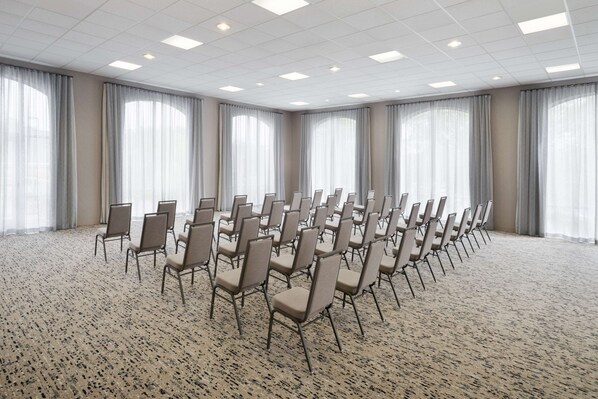 Un salón de conferencias con filas de sillas, ventanas amplias con cortinas translúcidas y una alfombra con manchas.