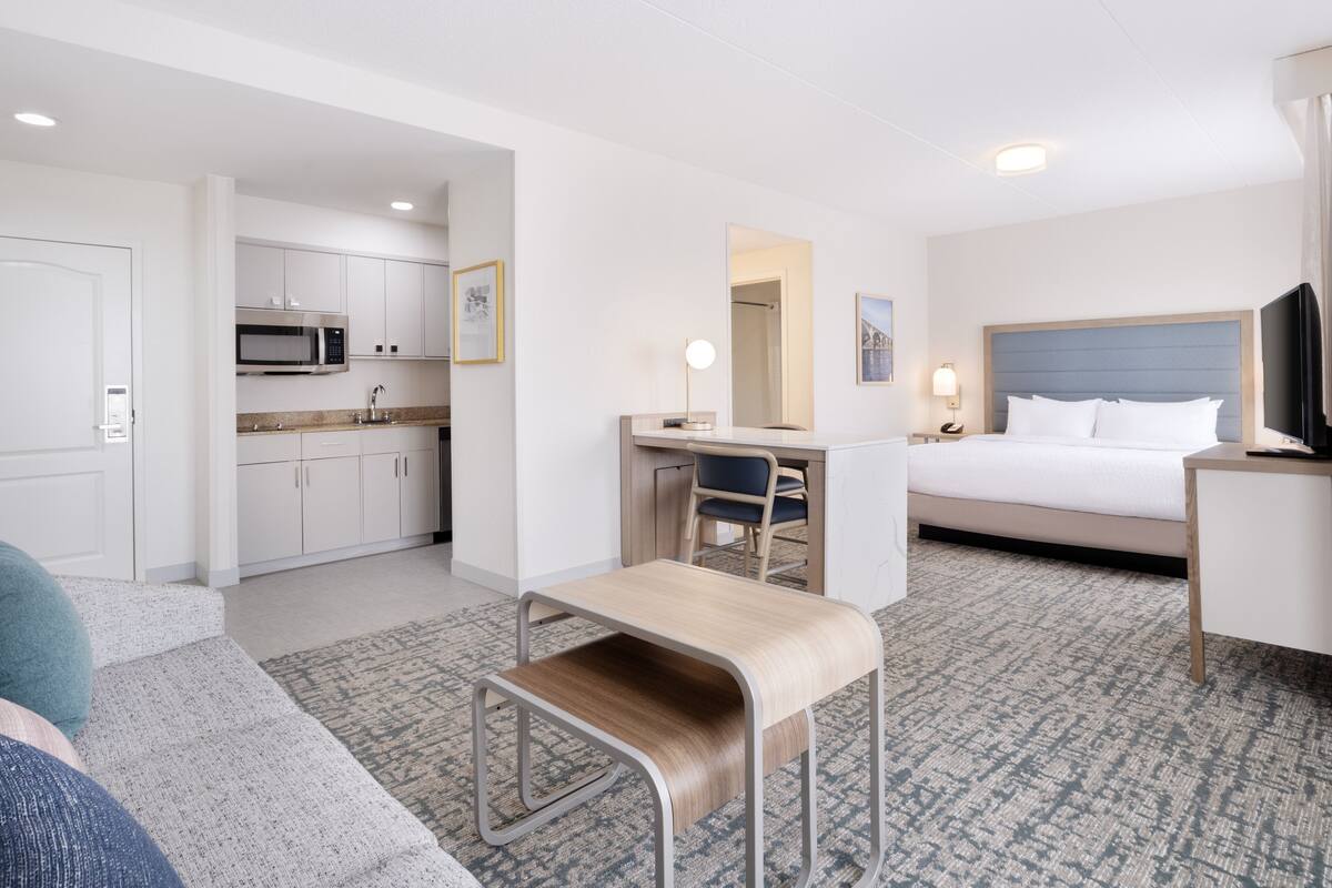 A hotel room with a bed, desk, chair, and kitchenette.