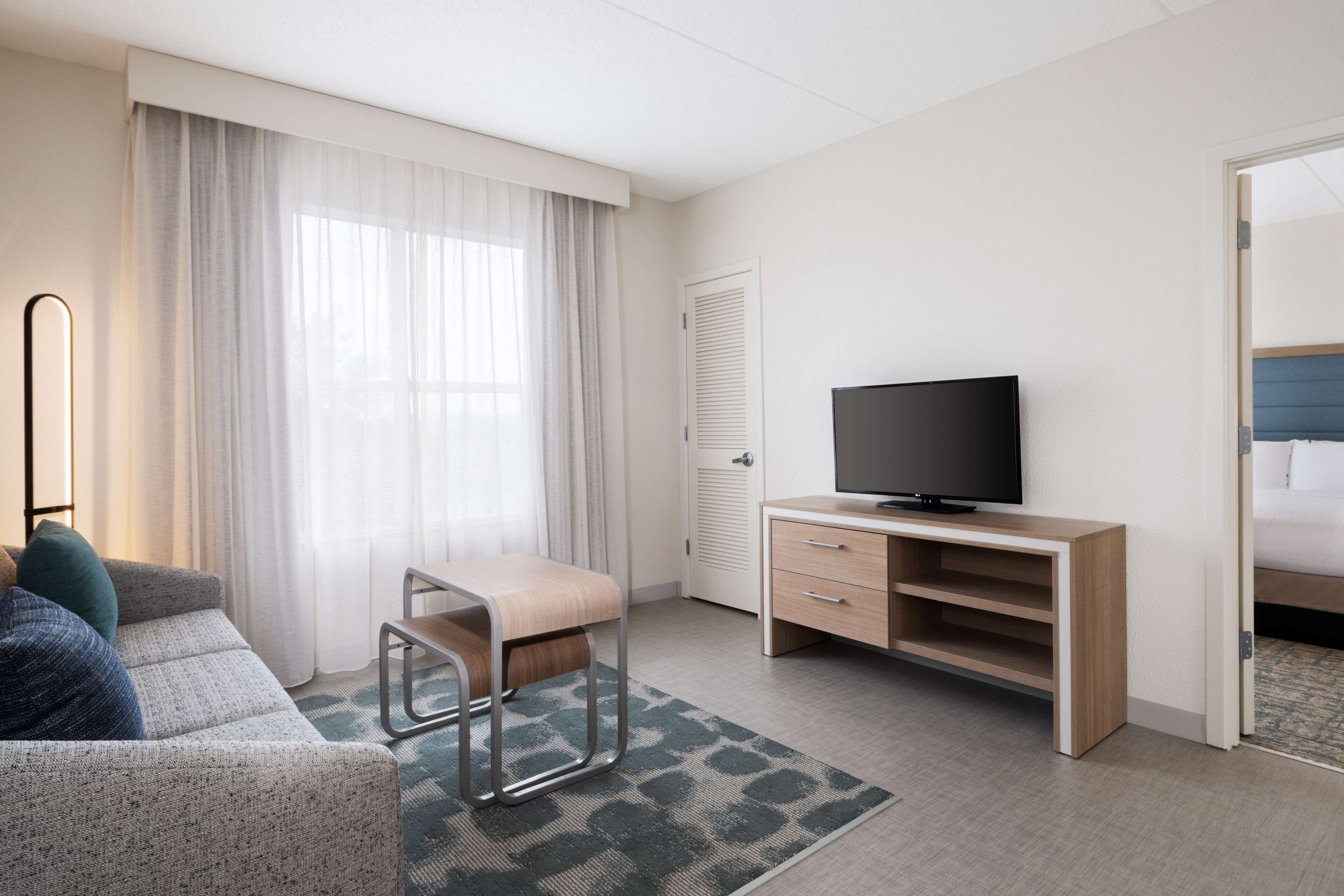 Ett hotellrum med en soffa, en tv på ett träställ, ett skrivbord med lådor, en stol och en säng med en blå sänggavel.