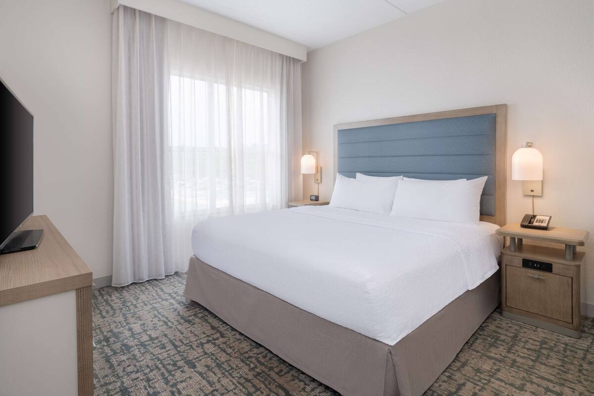 A hotel room with a large bed, a bedside table with a lamp, a TV on a stand, and a window with sheer curtains.
