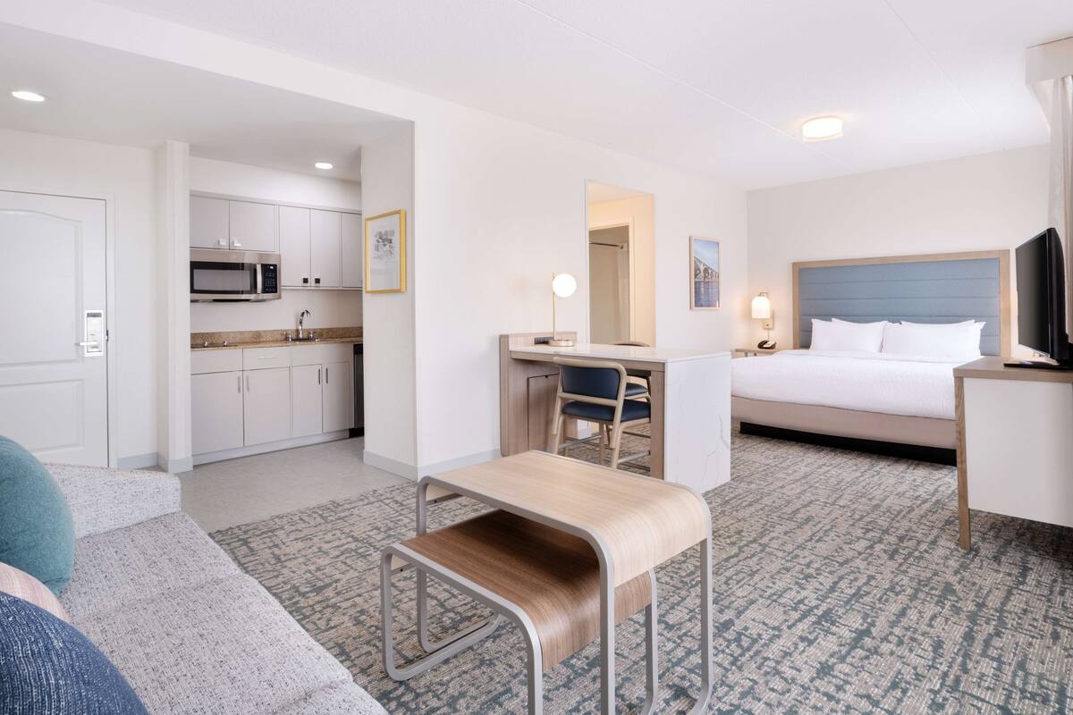 A hotel room with a bed, desk, chair, kitchenette, and a small bench.