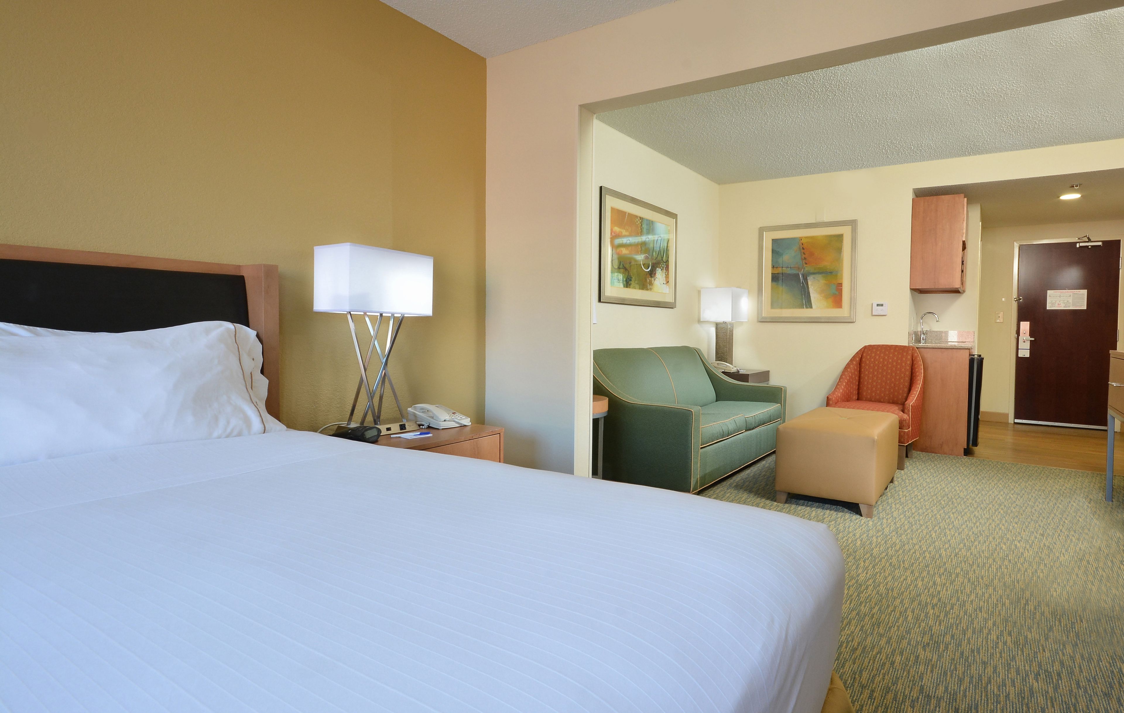 A hotel room with a large bed, a desk with a lamp, a green armchair, a red armchair, and a small ottoman.