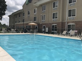 Zona de piscina hotelera con sillas de descanso y sombrillas, un cartel de prohibición de saltos y un edificio de ladrillo con numerosas ventanas.