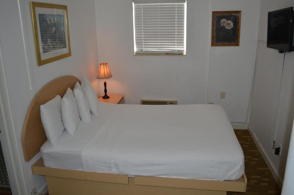 A single bed with a wooden headboard, a bedside lamp, a window with blinds, and a wall-mounted TV.