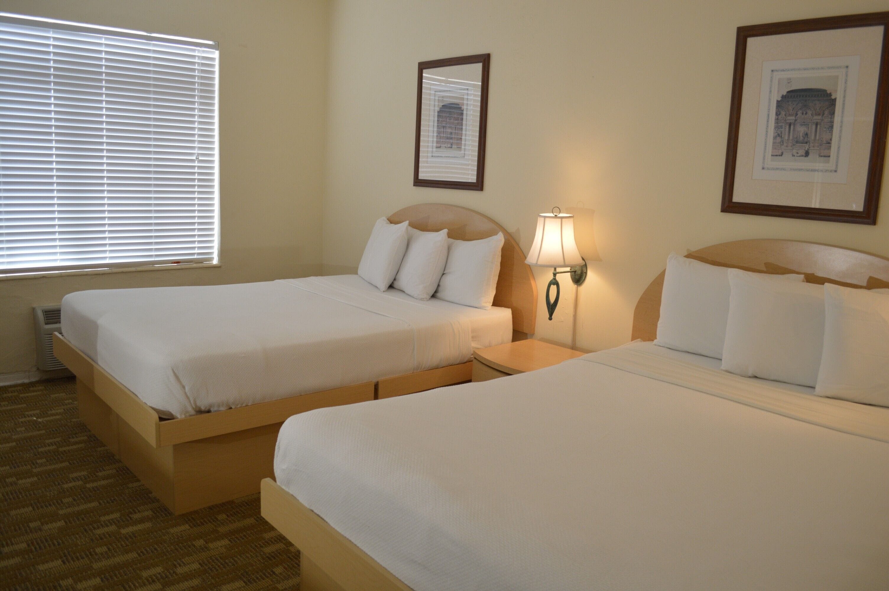 A hotel room with two single beds, a window with blinds, a wall-mounted lamp, and framed pictures.