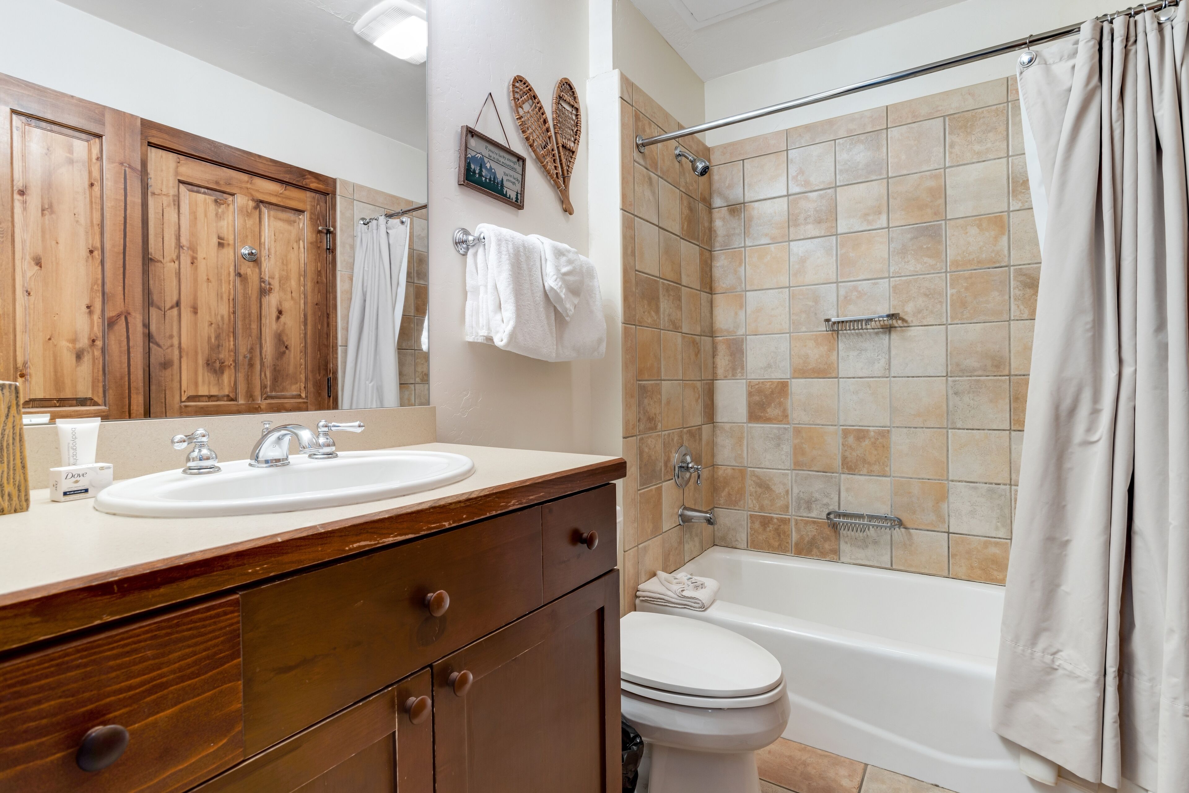 Un baño con tocador de madera, lavamanos, inodoro y ducha con baldosas color beige.
