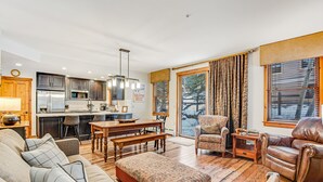 A spacious living area with a sofa, ottoman, and leather armchair. A wooden dining table with chairs is positioned near a kitchen with a refrigerator and microwave. Large windows provide a view of the outdoors. - Main Street Junction (Breckenridge)