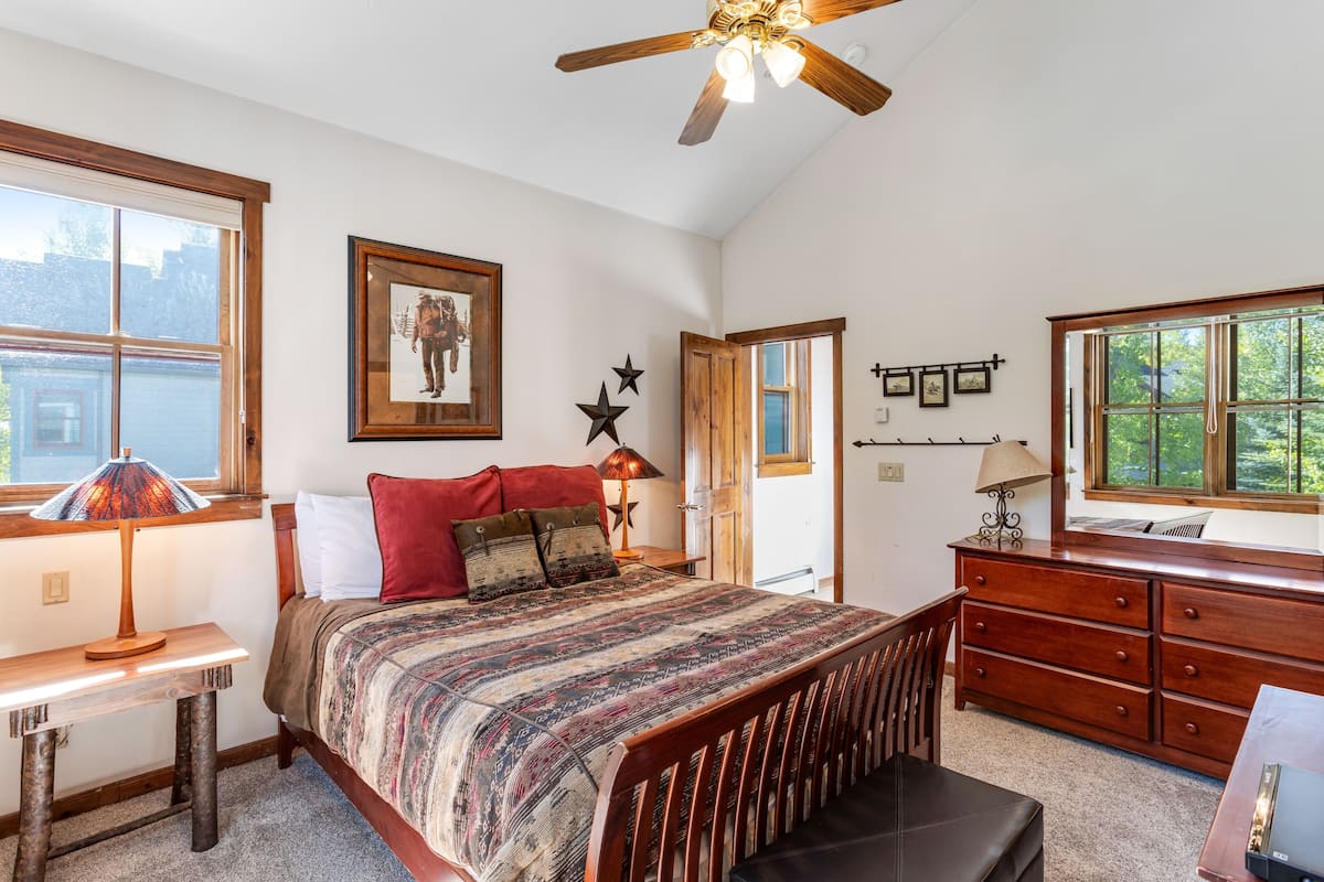 Une chambre à coucher comprenant un lit, une commode en bois, un bureau et un ventilateur de plafond.