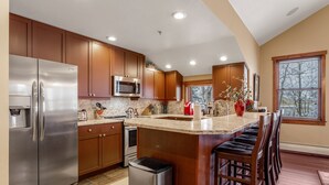A modern kitchen with stainless steel appliances, wooden cabinets, a marble countertop, and a bar area with stools. - Main Street Junction (Breckenridge)