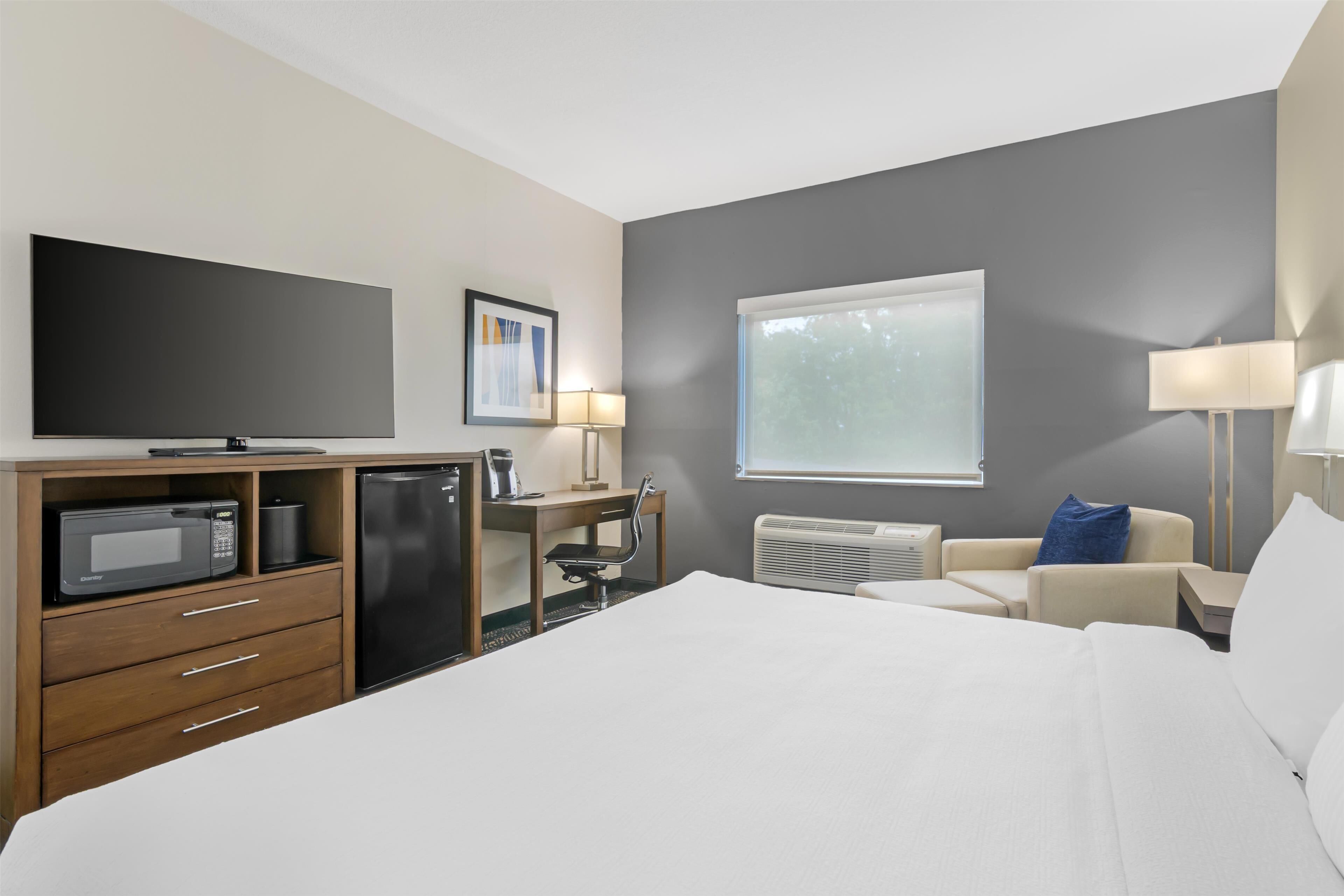 A hotel room with a bed, a desk with a chair, a microwave, a refrigerator, a TV, a window, and a lamp.
