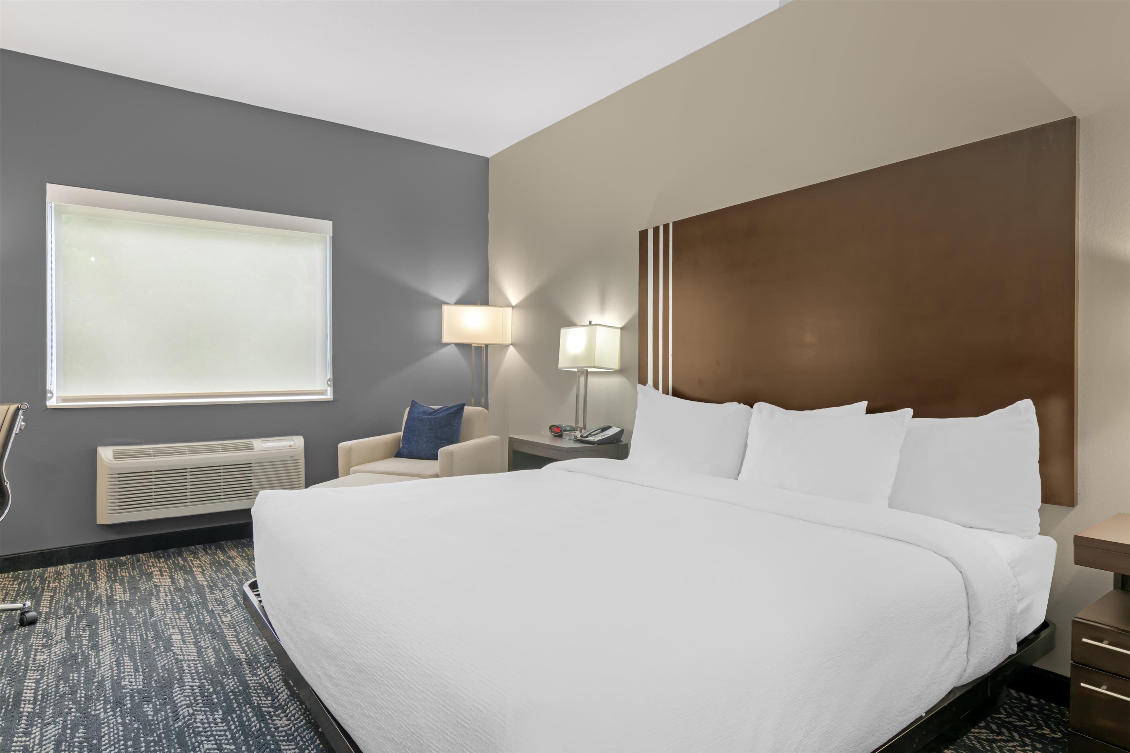 A hotel room with a large bed, a chair, a desk with a lamp, a window, and a wall-mounted heater.