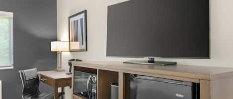 A hotel room with a desk, chair, television, and a Haier refrigerator.