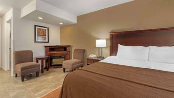A hotel room with a large bed, two armchairs, a small table, a lamp, and a fireplace.