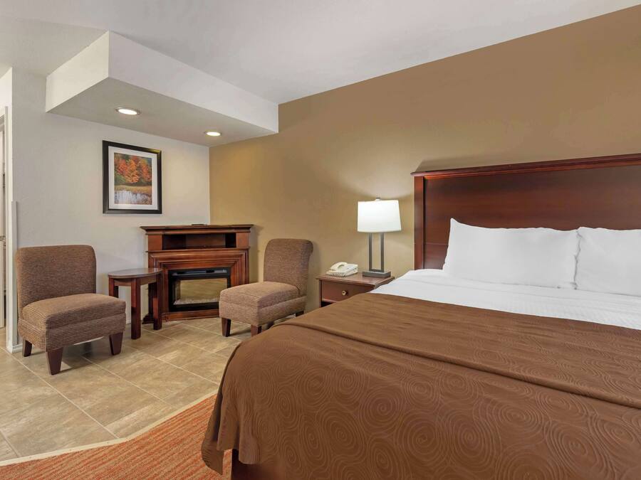 A hotel room with a large bed, two armchairs, a small table, a lamp, and a fireplace.