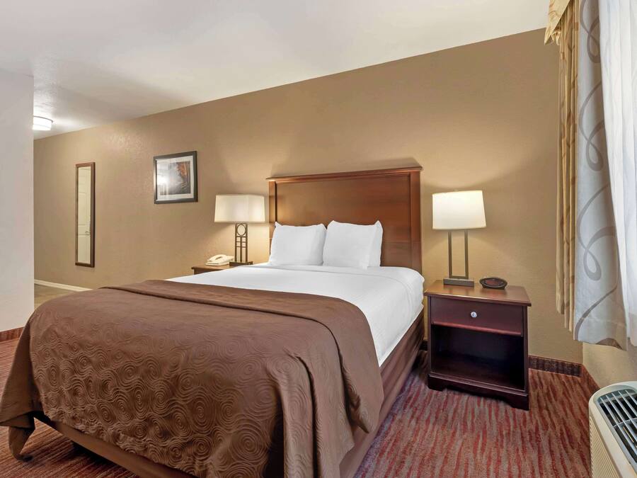 A hotel room with a large bed, two bedside tables with lamps, a nightstand, a wall-mounted telephone, and a window with curtains.