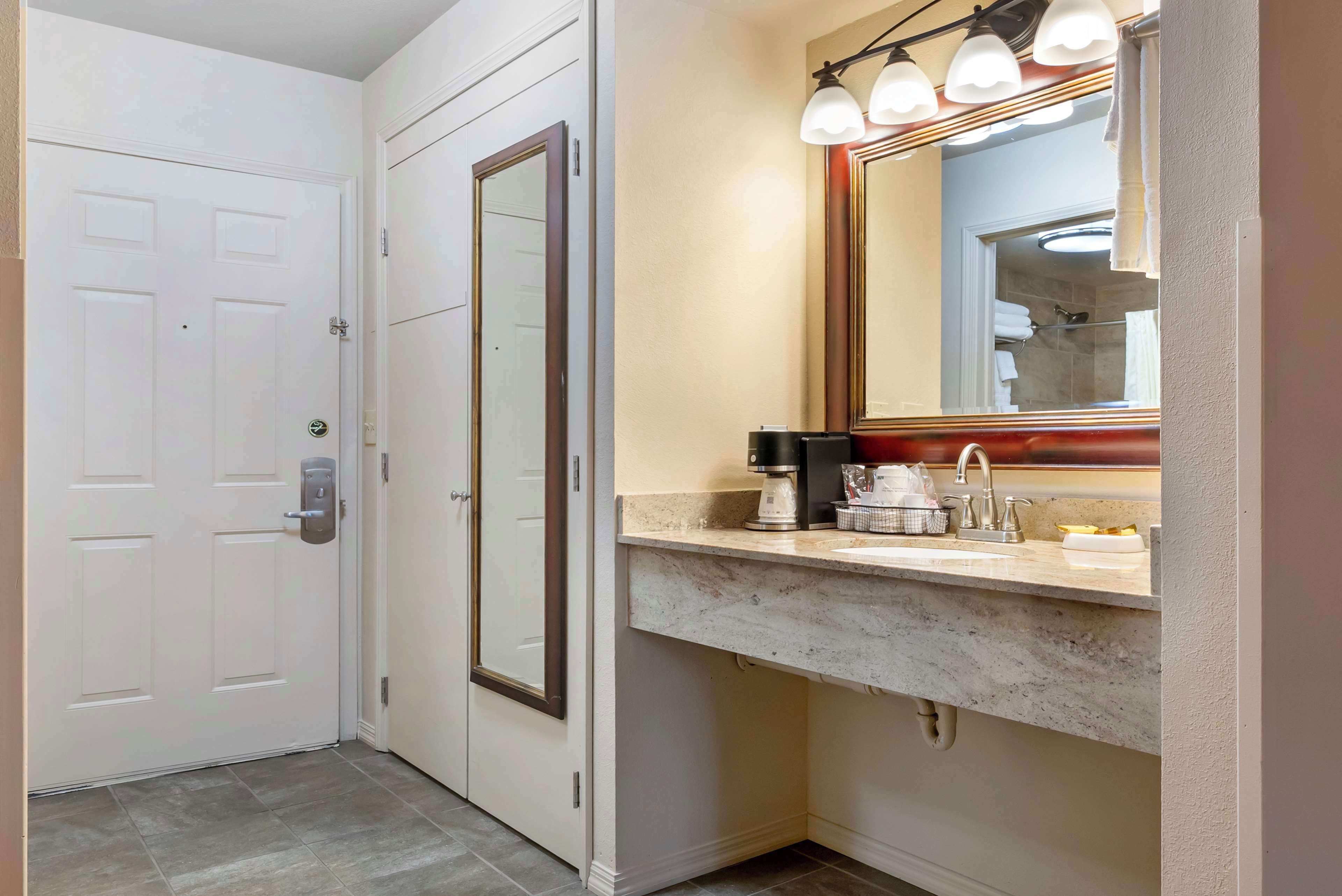 A bathroom with a white door, a mirror with lights, a sink, and a coffee maker.
