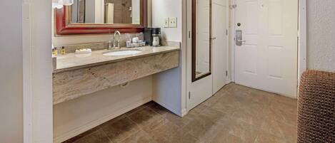 A bathroom with a marble sink, a large mirror with lights, a white door, and a wicker bench.