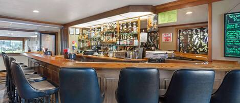 A bar area with a wooden counter, black bar stools, and a variety of liquor bottles on shelves.