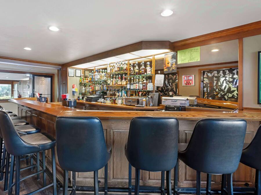 Un espace bar avec un comptoir en bois, des tabourets de bar noirs et une variété de bouteilles d’alcool exposées sur des étagères.