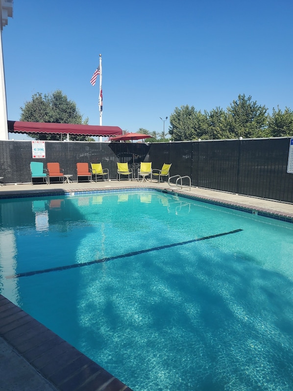 A clear swimming pool with a flag, a poolside area with chairs, and a fence.