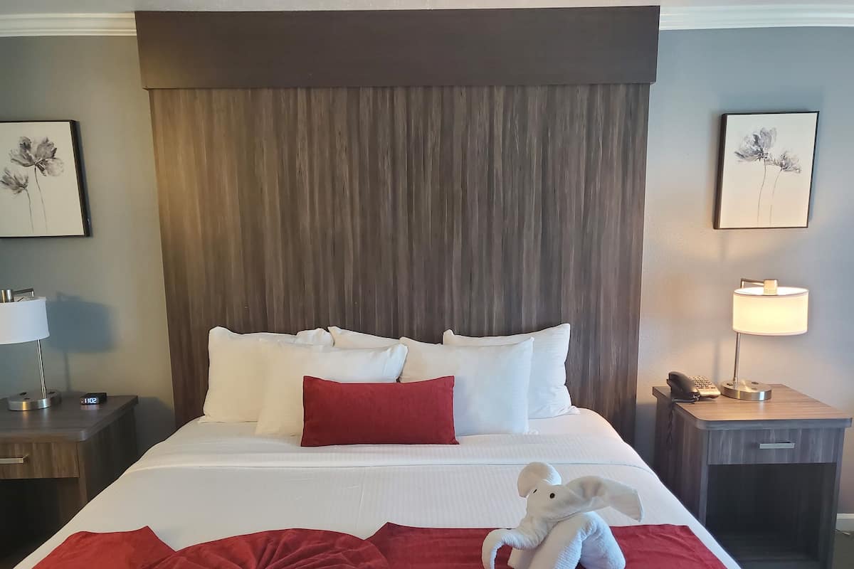 Una cama bien hecha con ropa de cama blanca y roja, una almohada roja y una almohada con forma de elefante blanco. Hay dos mesitas de noche, cada una con una lámpara y un teléfono. La habitación tiene un cabecero de madera y una pared gris.