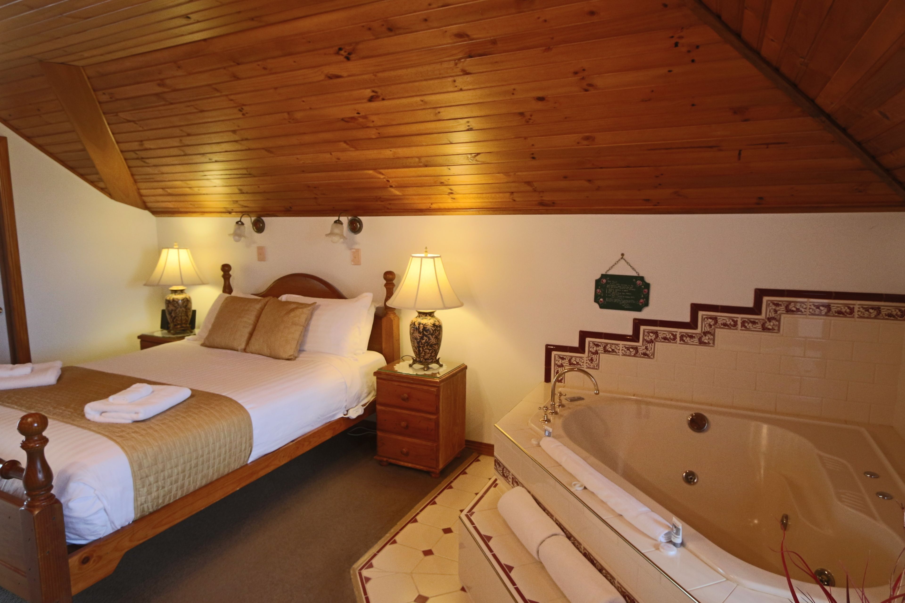 A bedroom with a wooden ceiling, a large bed, two bedside lamps, a nightstand, a dresser, a bathroom with a bathtub, and a wall-mounted step ladder.