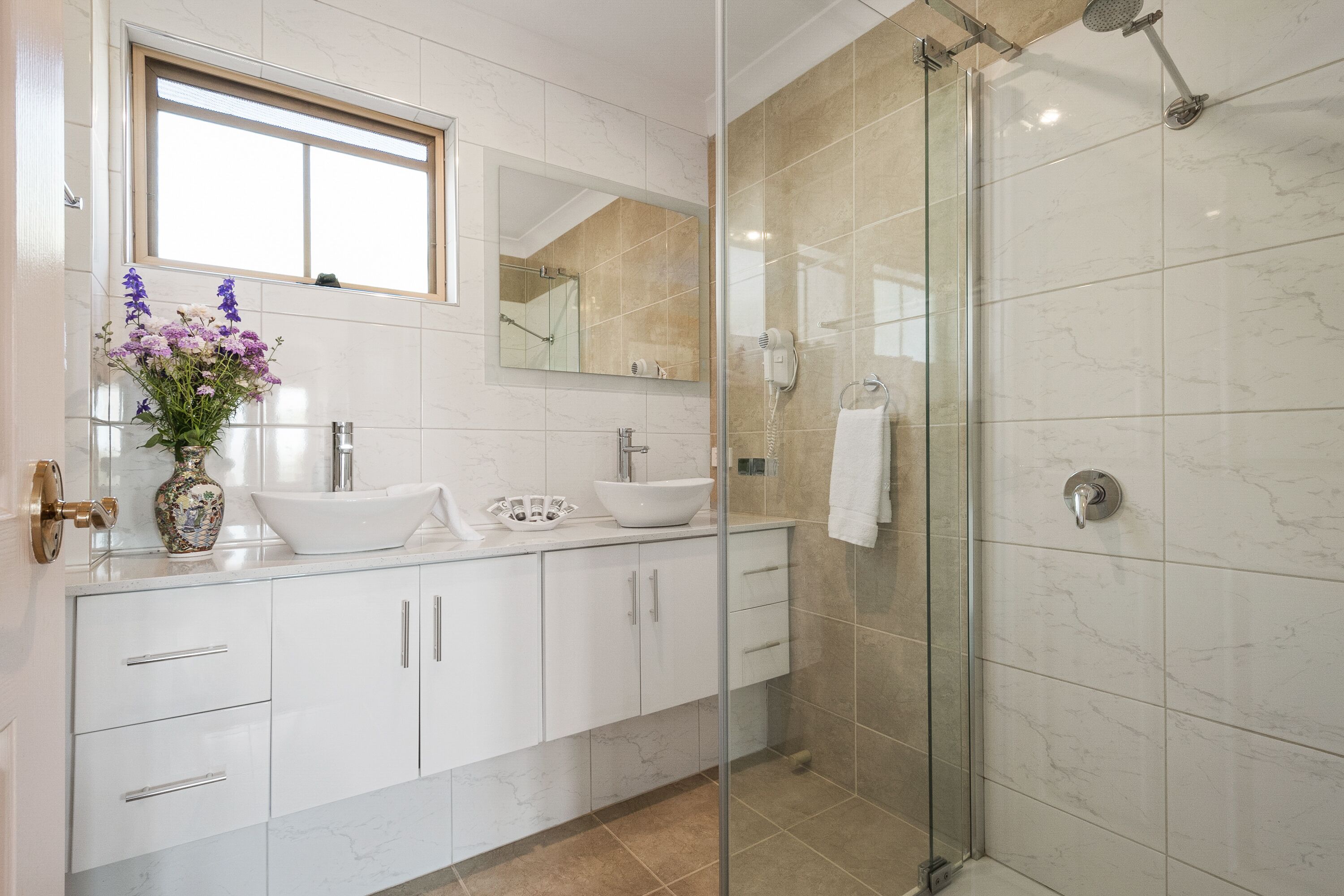A modern bathroom with a glass-enclosed shower, a large mirror, and a vase of flowers on the vanity.