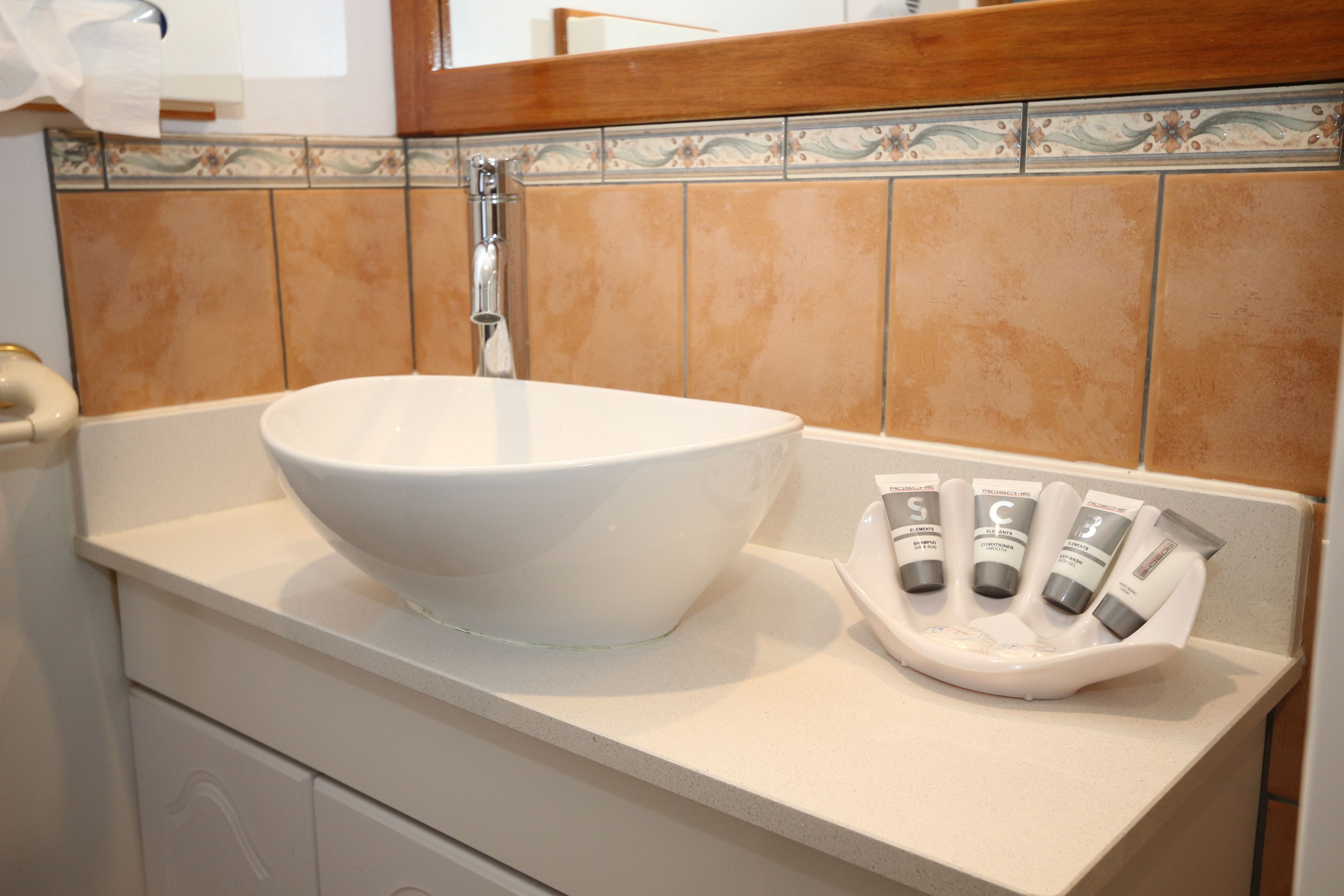 Un baño con un lavamanos grande de color blanco, un espejo y un jabonera con varios tubos.