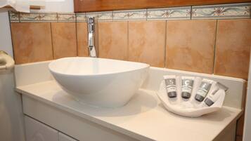 Un baño con un lavamanos grande de color blanco, un espejo y un jabonera con varios tubos.
