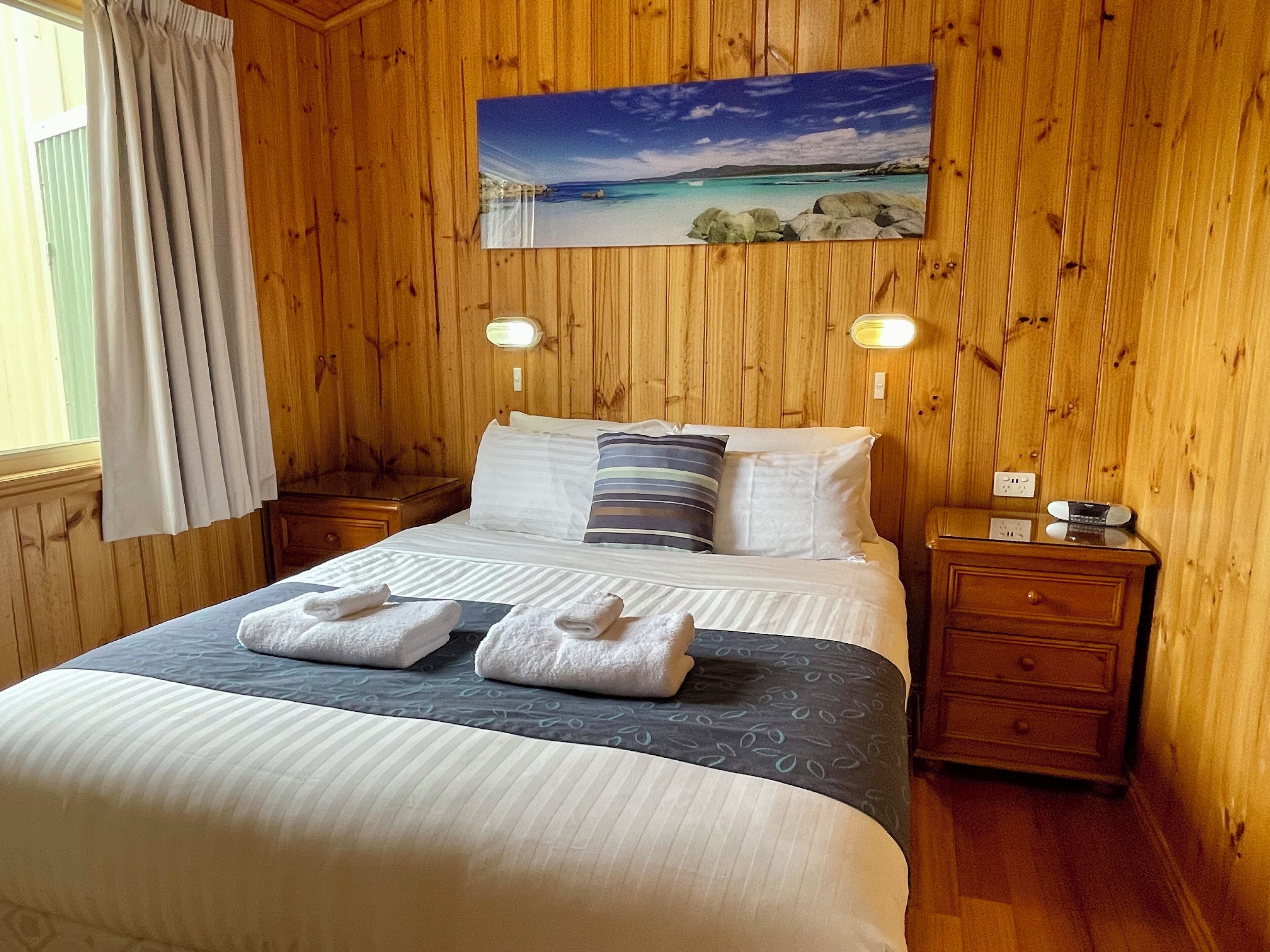 A wooden-paneled room with a bed, two bedside tables, a painting of a beach, and a window with curtains.