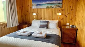 A wooden-paneled room with a bed, two bedside tables, a painting of a beach, and a window with curtains.
