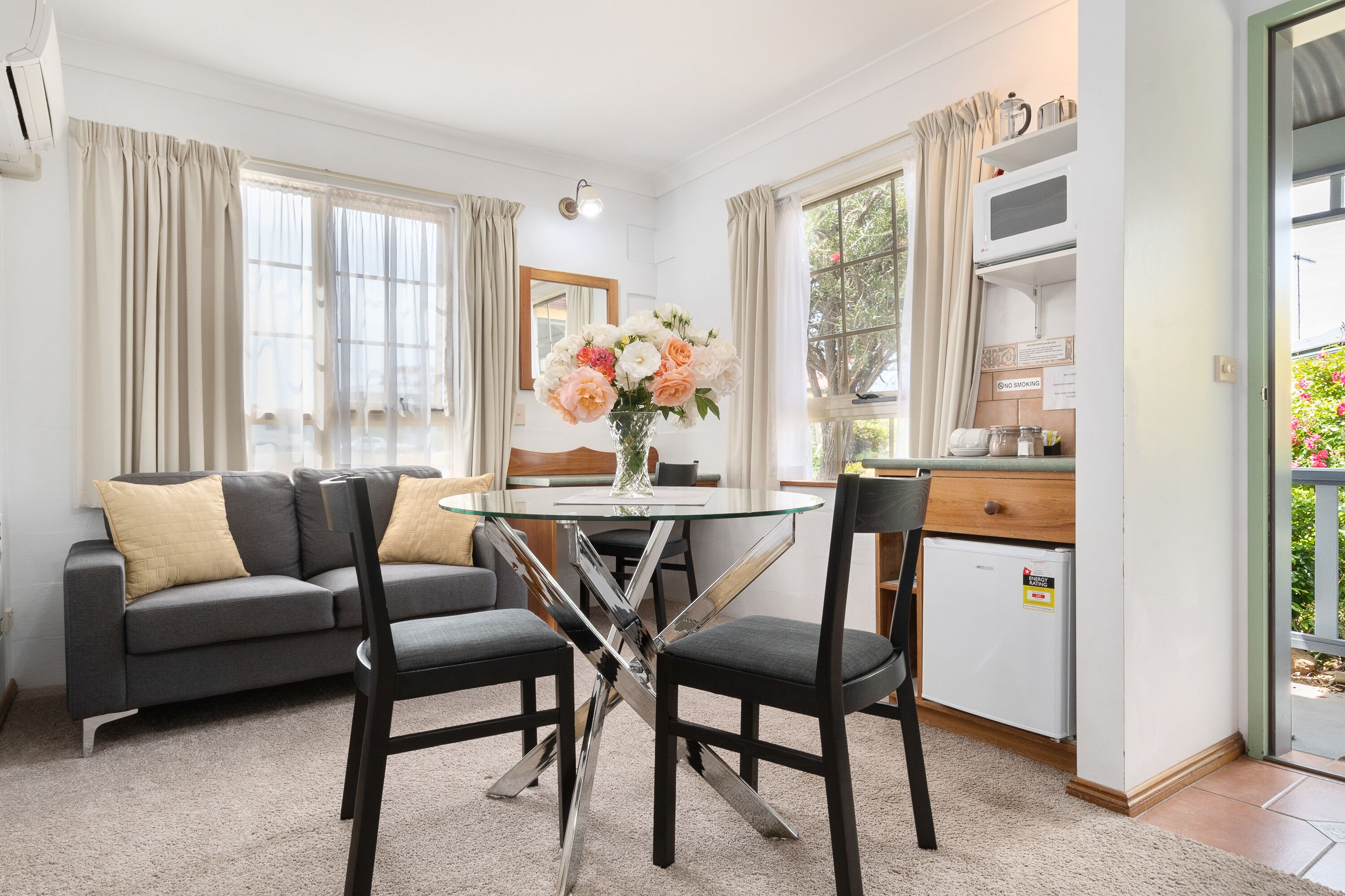 A modern living room with a glass-top dining table, two black chairs, a grey sofa with beige cushions, and a vase of flowers.