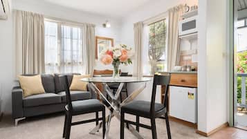 A modern living room with a glass-top dining table, two black chairs, a grey sofa with beige cushions, and a vase of flowers.
