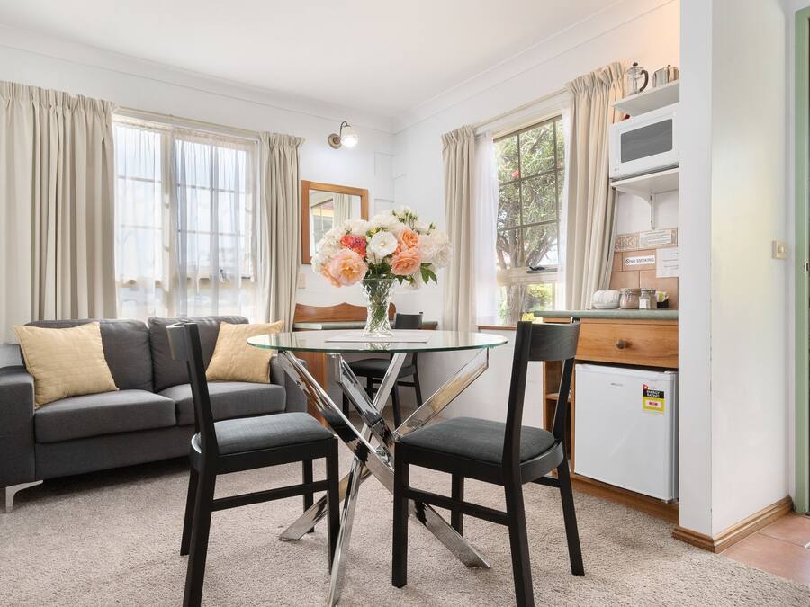 A modern living room with a glass-top dining table, two black chairs, a grey sofa with beige cushions, and a vase of flowers.