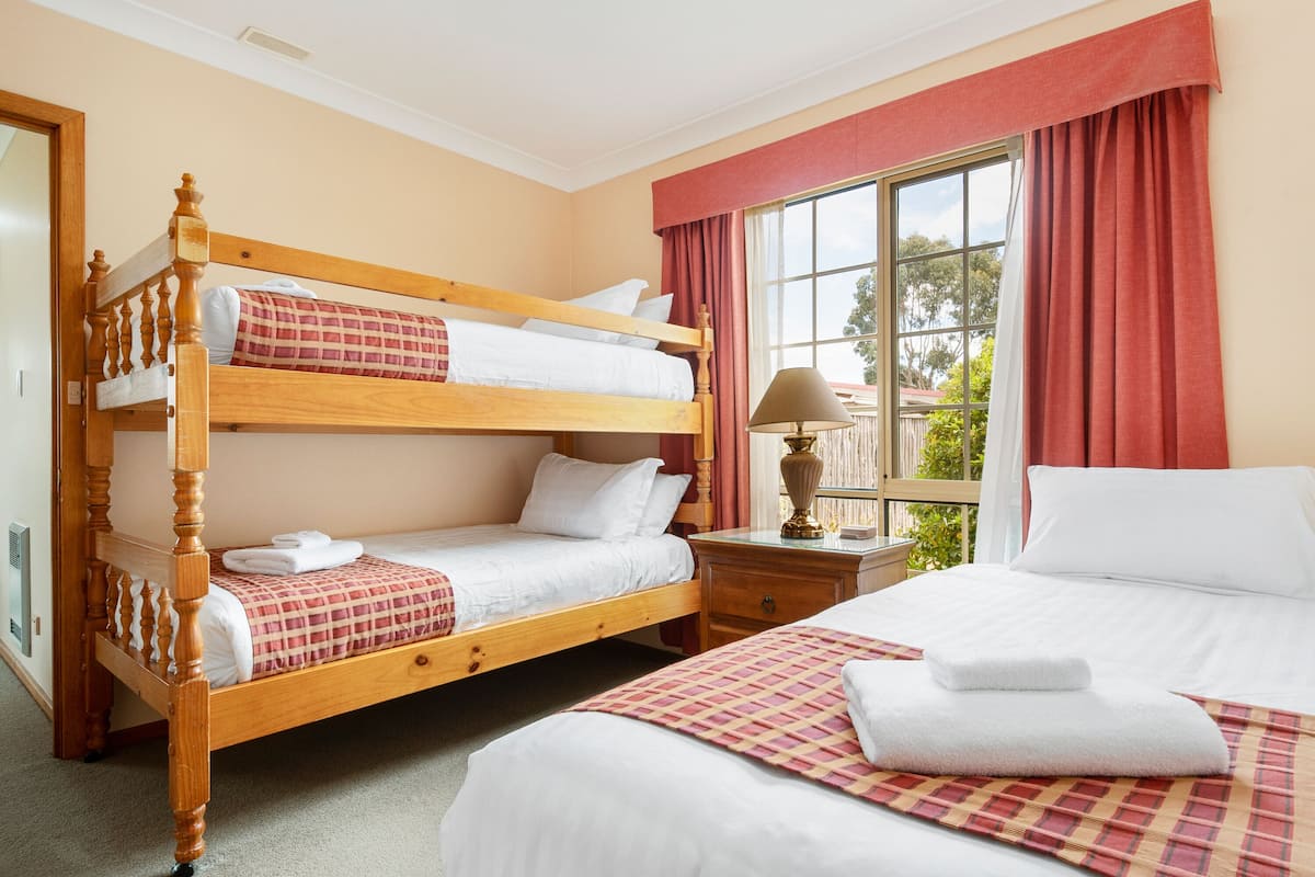 Una habitación de hotel con una cama litera de madera, una cama individual, una mesita de noche con lámpara y una ventana con cortinas.