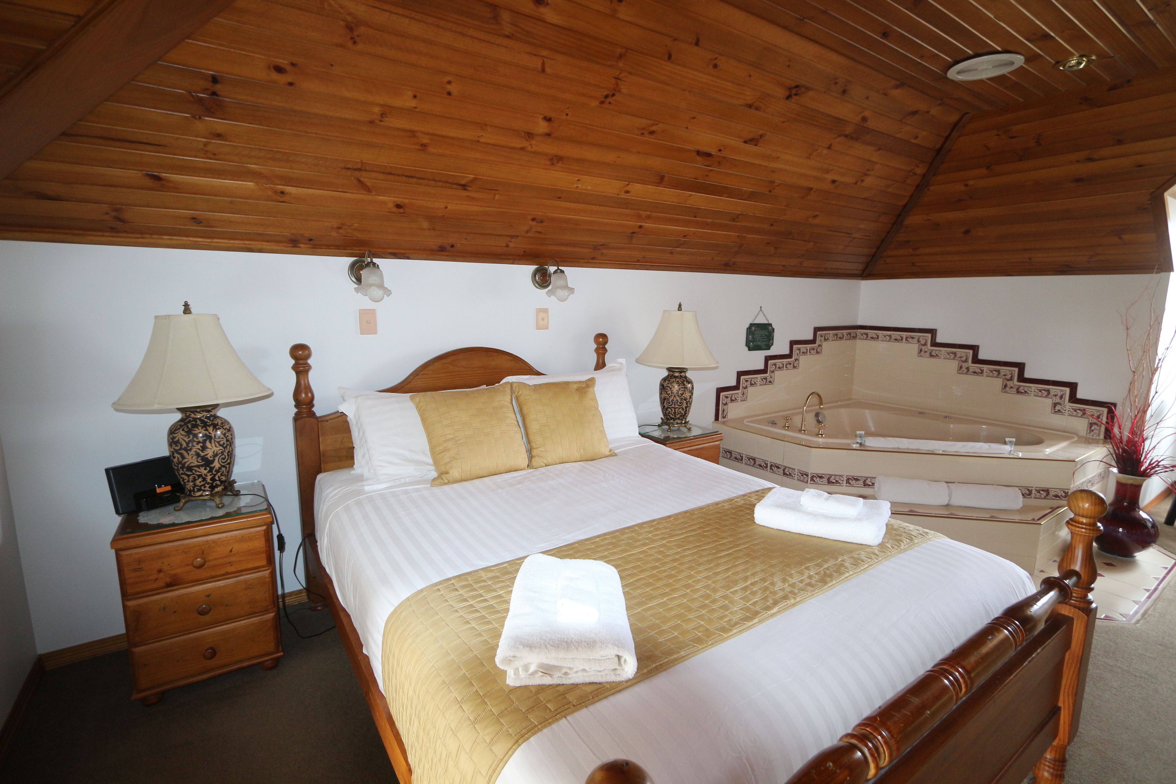 Een slaapkamer met een houten plafond, een groot bed met een gouden sprei, twee nachtlampjes, een nachtkastje, een houten dressoir, en een badkamer met een bad en een wastafel.
