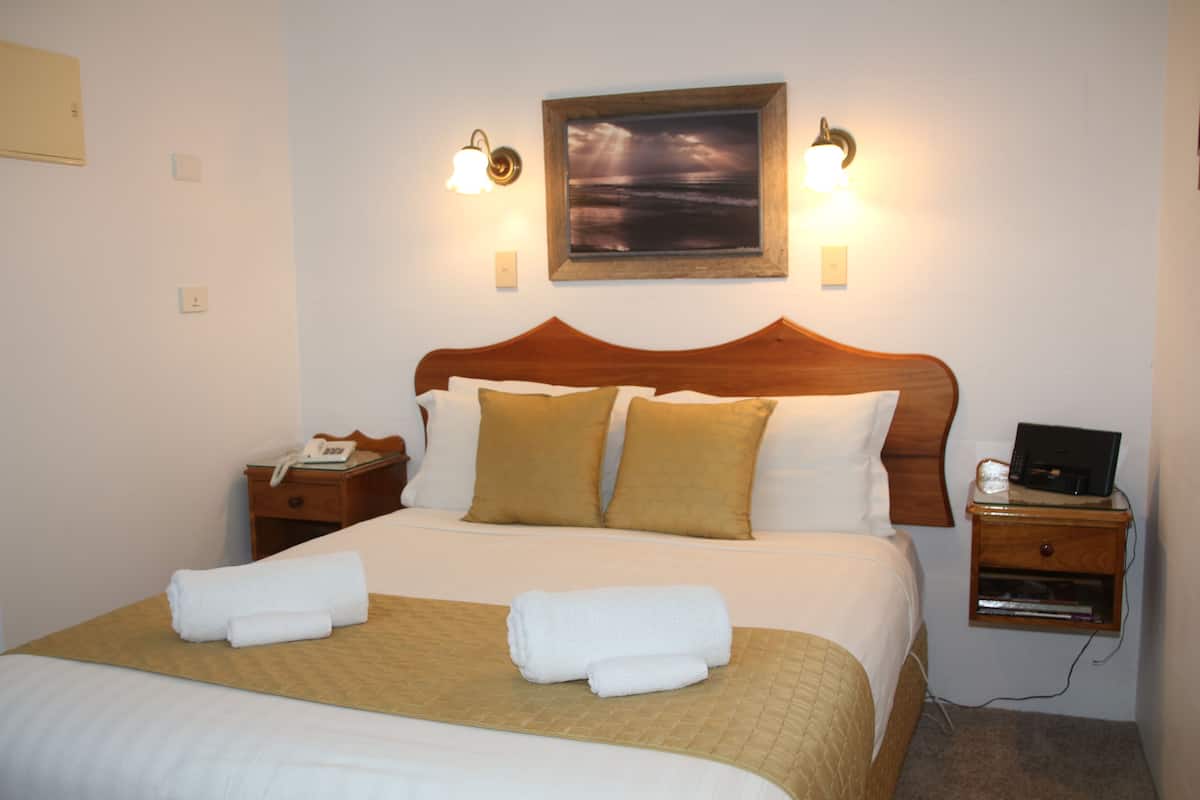 Una cama bien hecha con dos almohadas, una mesita de noche con lámpara, un cuadro enmarcado en la pared y un mueble con cajones.