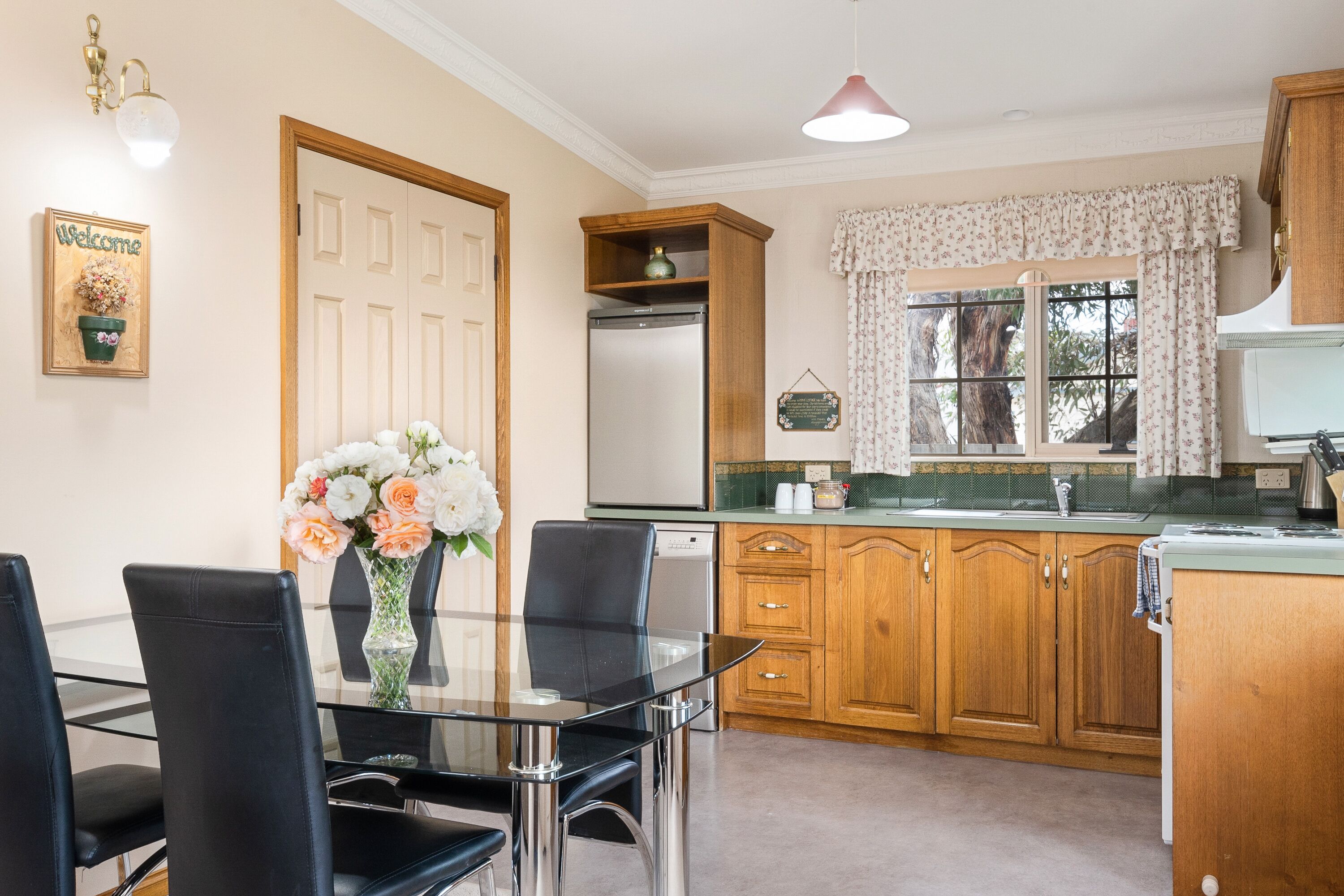 A kitchen with wooden cabinets, a stainless steel refrigerator, a glass dining table with chairs, and a window with floral curtains.