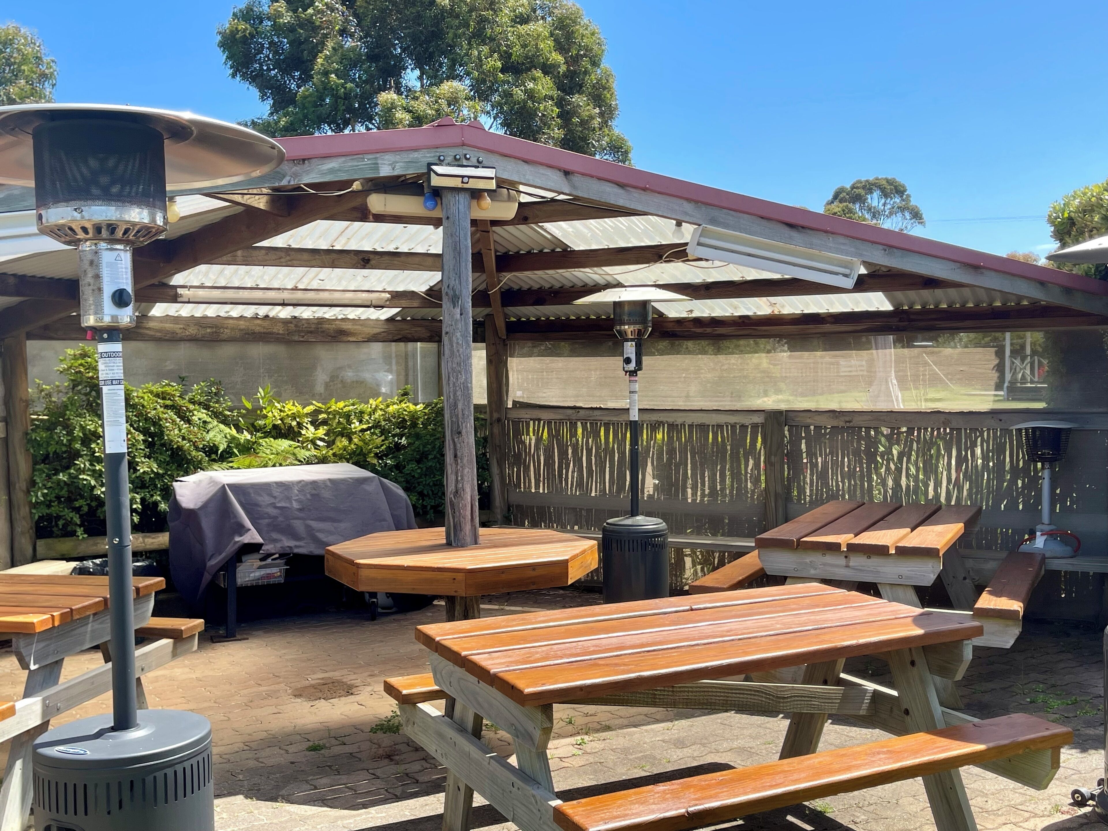 Un espace extérieur aménagé en terrasse, avec des tables et des bancs en bois, un coin salon abrité et un radiateur.