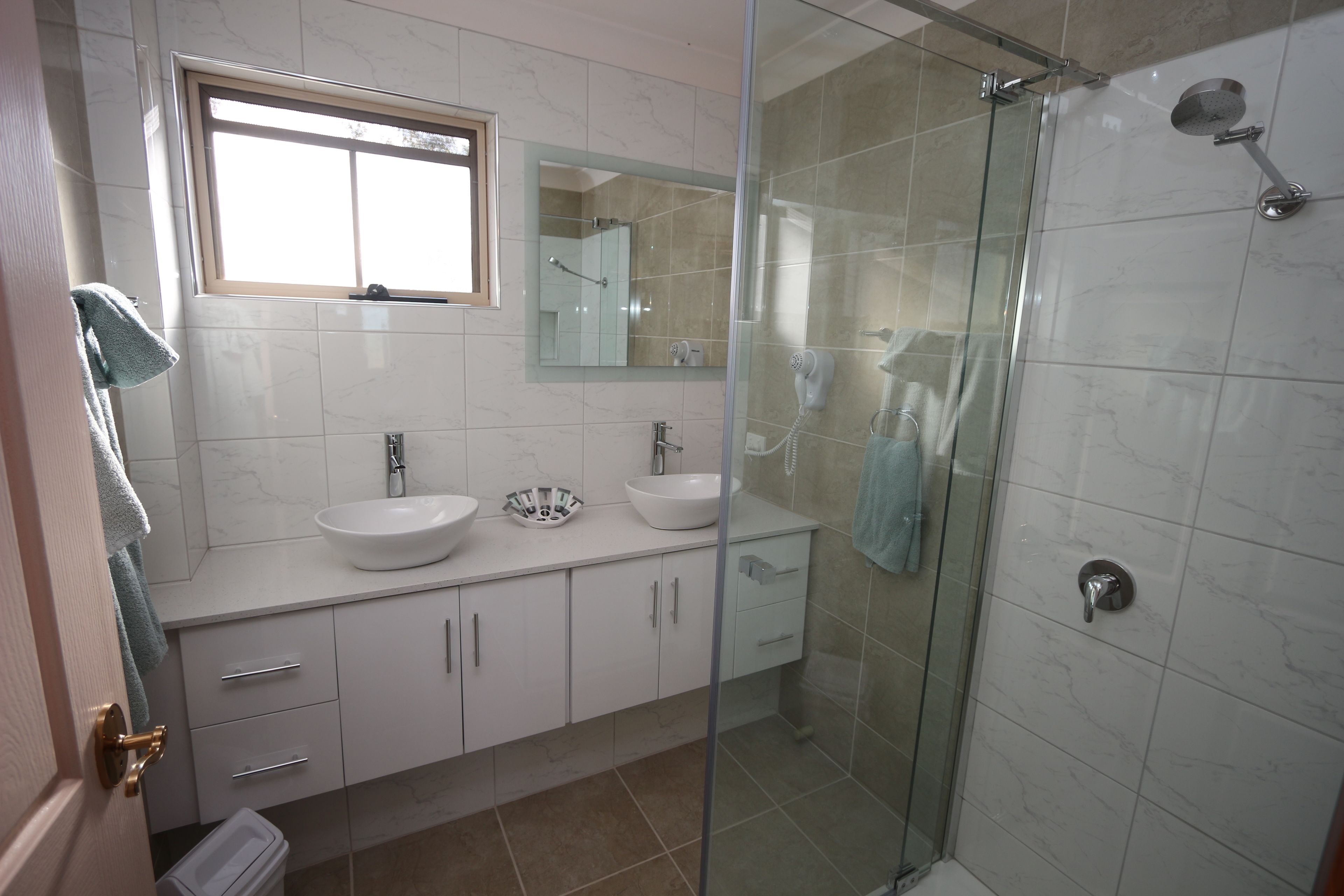 Een moderne badkamer met twee wastafels, een douchecabine van glas en een raam boven de wastafel.