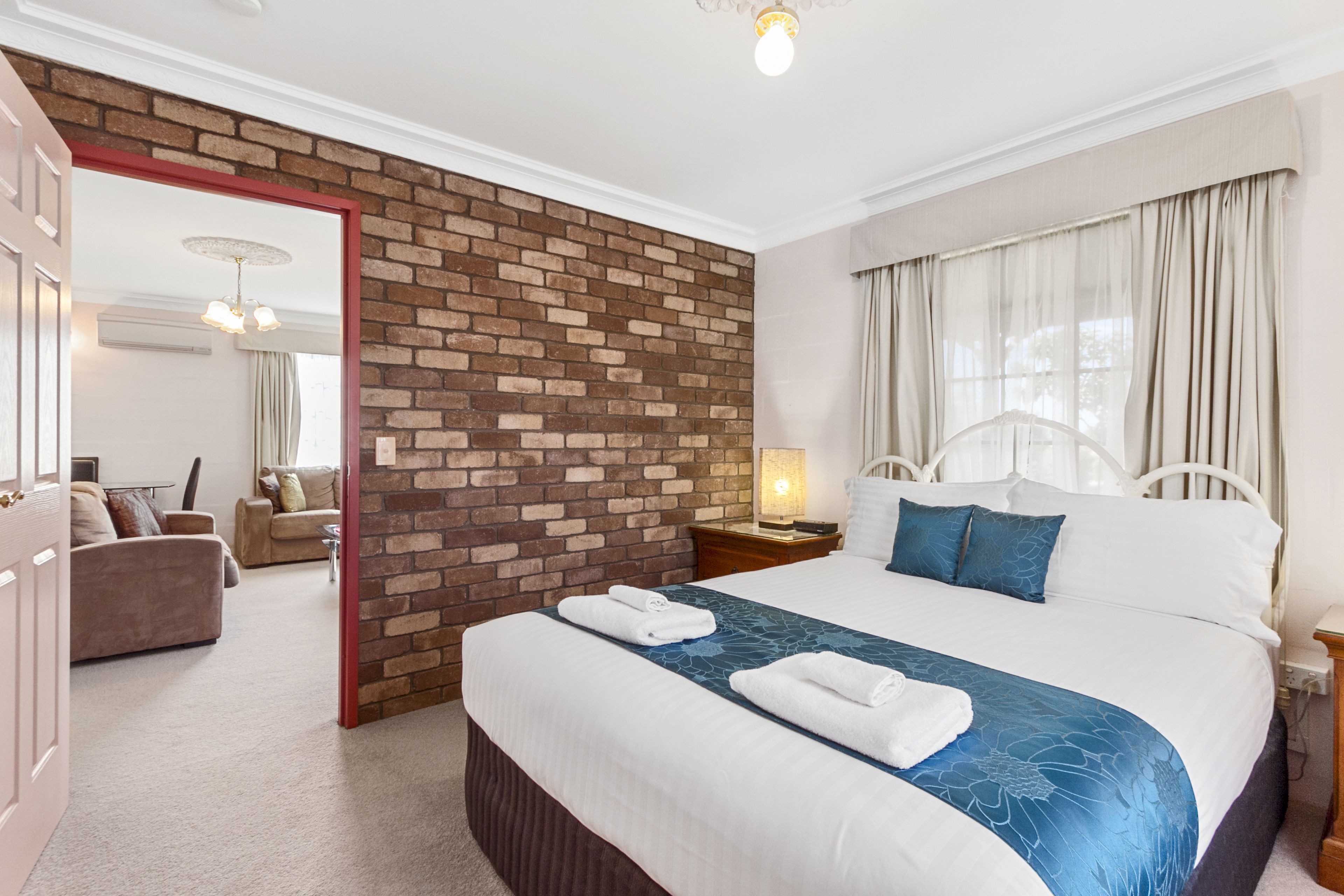 Quarto de hotel com parede de destaque em tijolos, uma cama grande com roupa de cama azul, uma mesa de cabeceira com abajur e vista para a área de estar através de uma porta aberta.