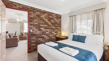 Quarto de hotel com parede de destaque em tijolos, uma cama grande com roupa de cama azul, uma mesa de cabeceira com abajur e vista para a área de estar através de uma porta aberta.