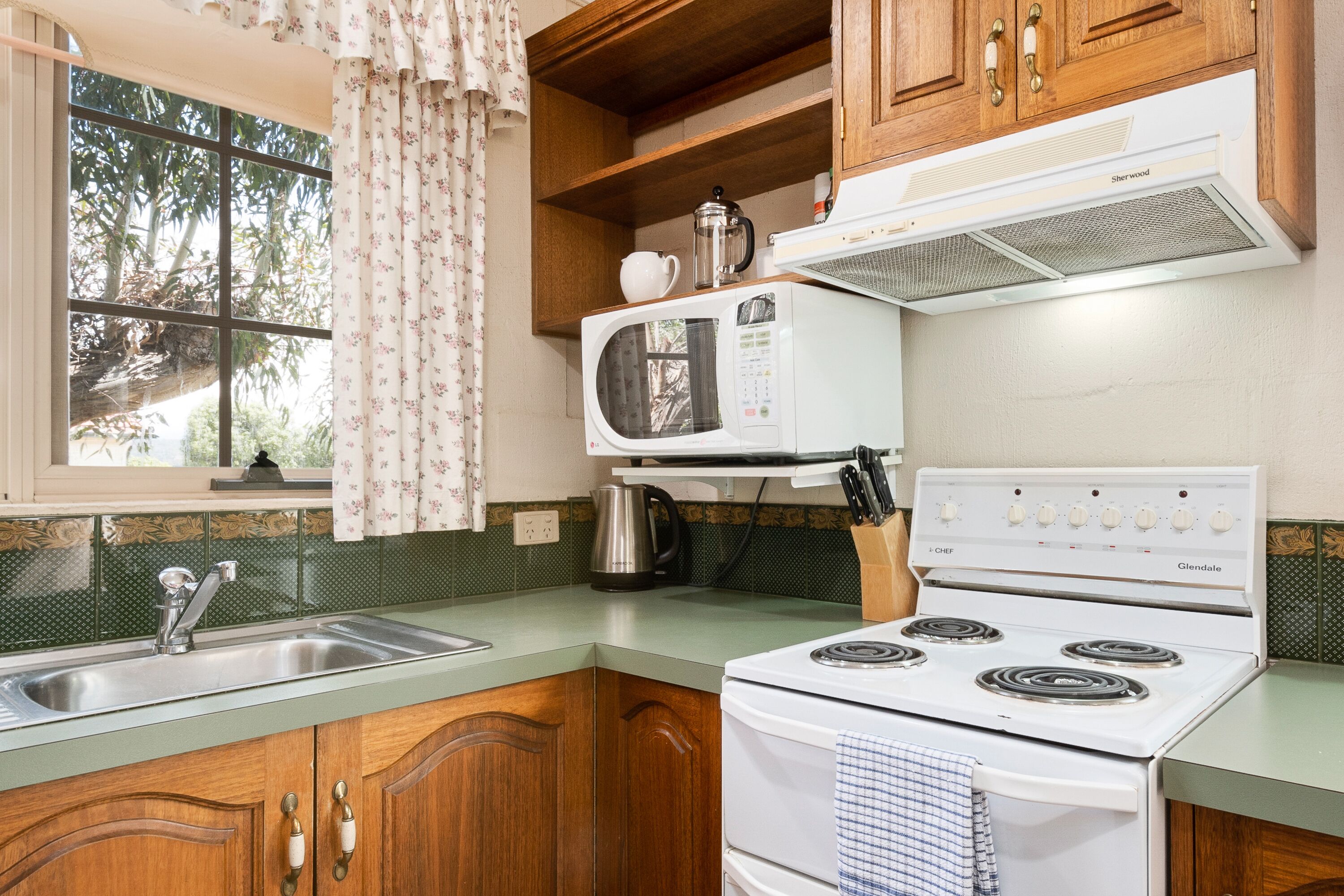 A kitchen with wooden cabinets, a white stove, a microwave, and a window with curtains.