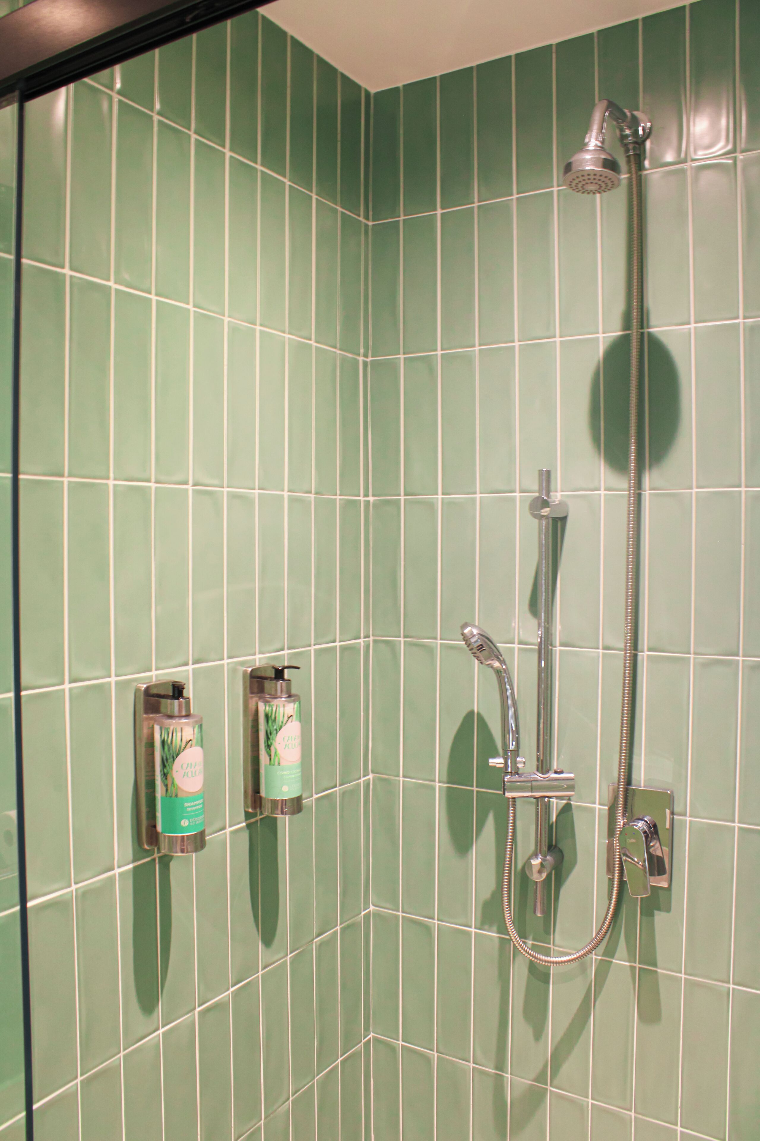 A shower area with green tiles, a handheld showerhead, and two soap dispensers on the wall.