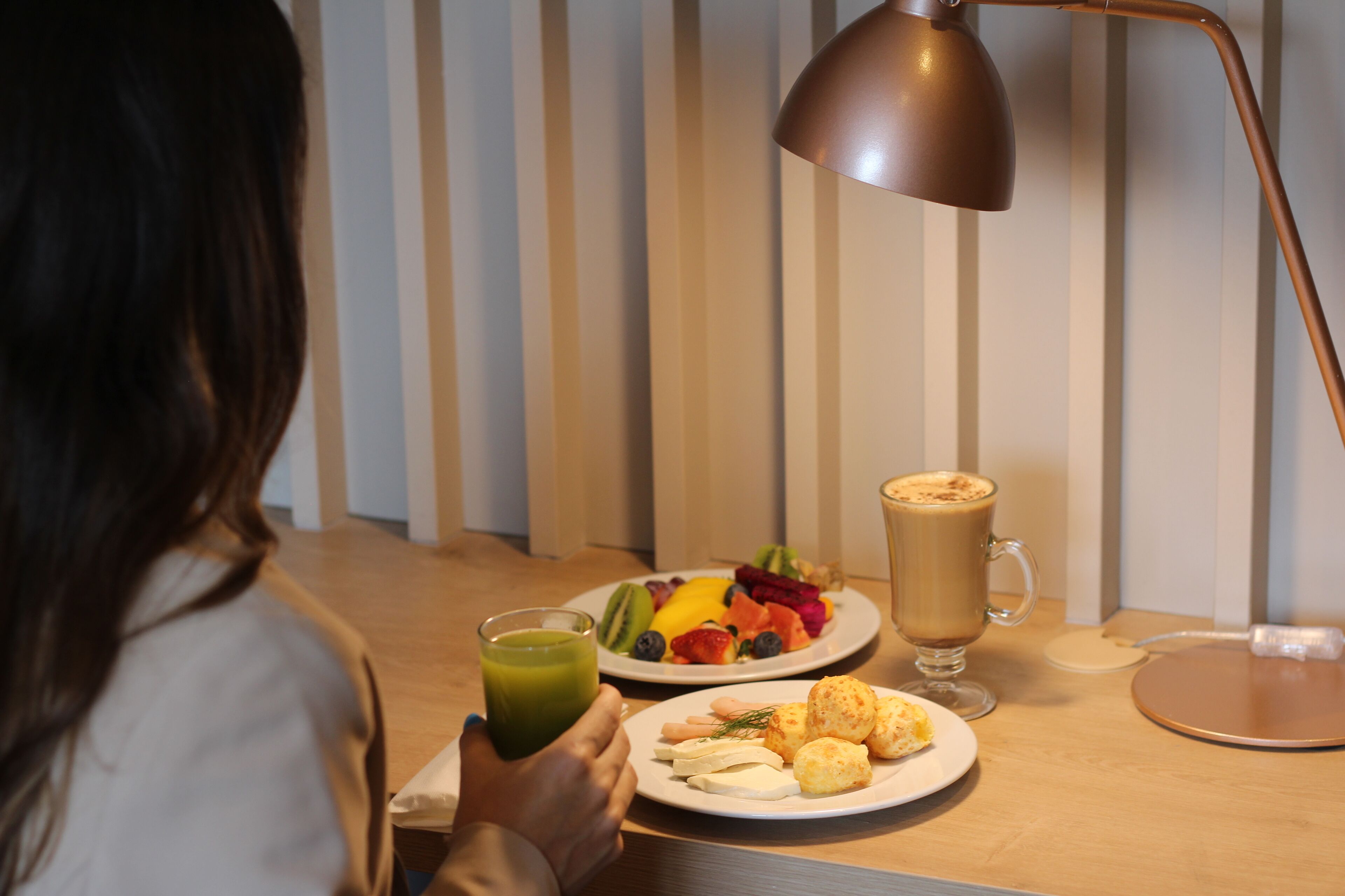 A person sitting at a table with a meal consisting of fruit, pastries, and a beverage, with a lamp and a charging device on the table.