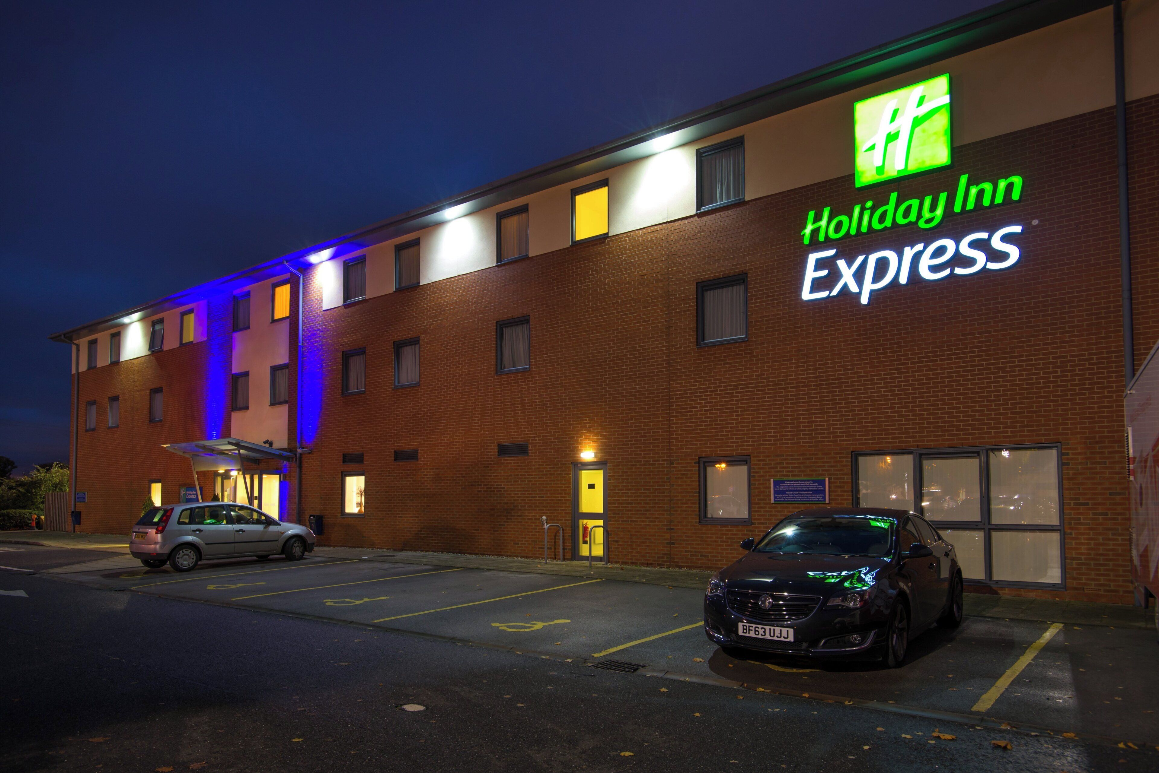 Edificio de Holiday Inn Express con un estacionamiento y dos coches aparcados en la entrada.
