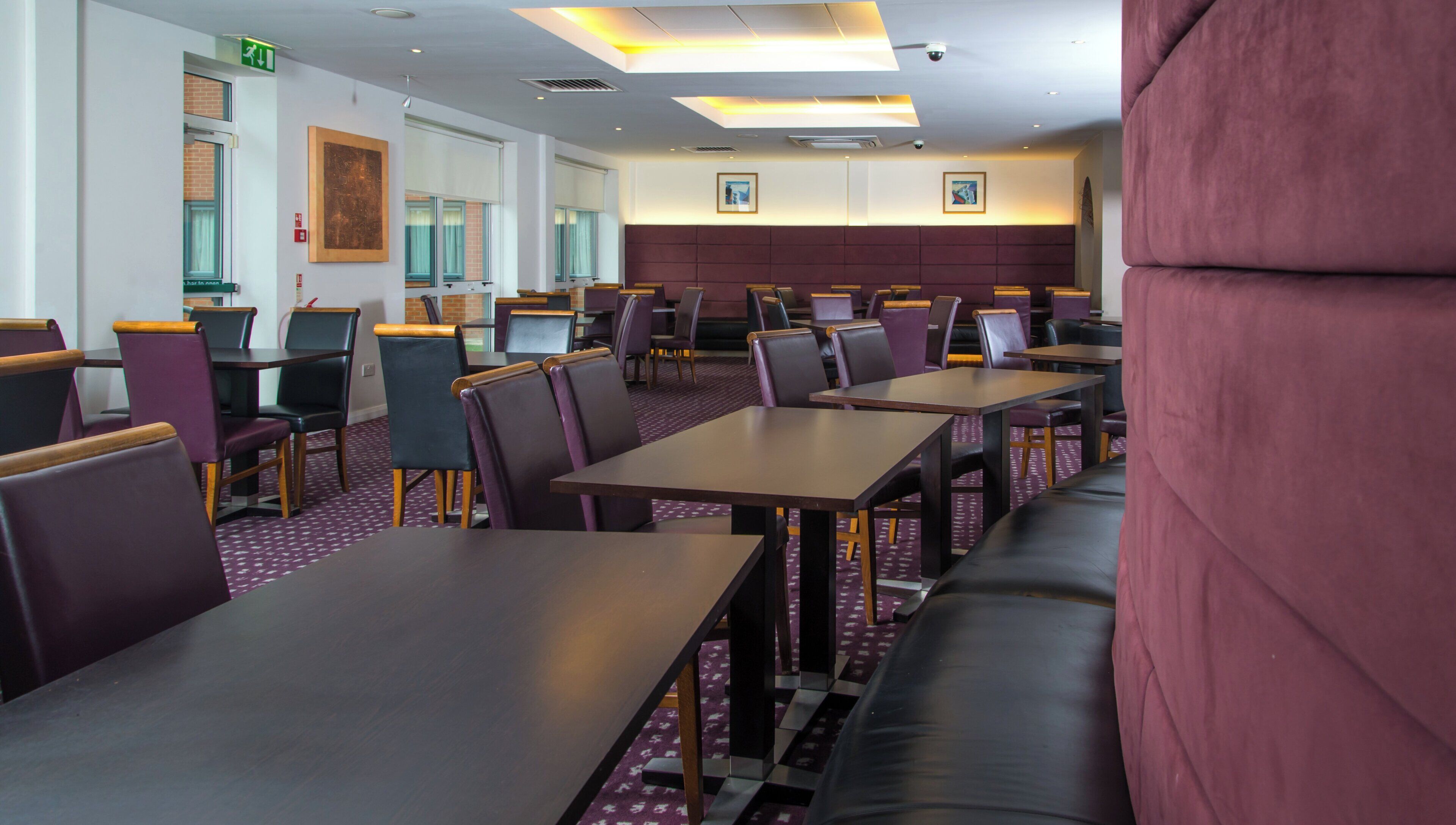 A hotel dining area with multiple tables and chairs, a patterned carpet, and a large cushioned bench.