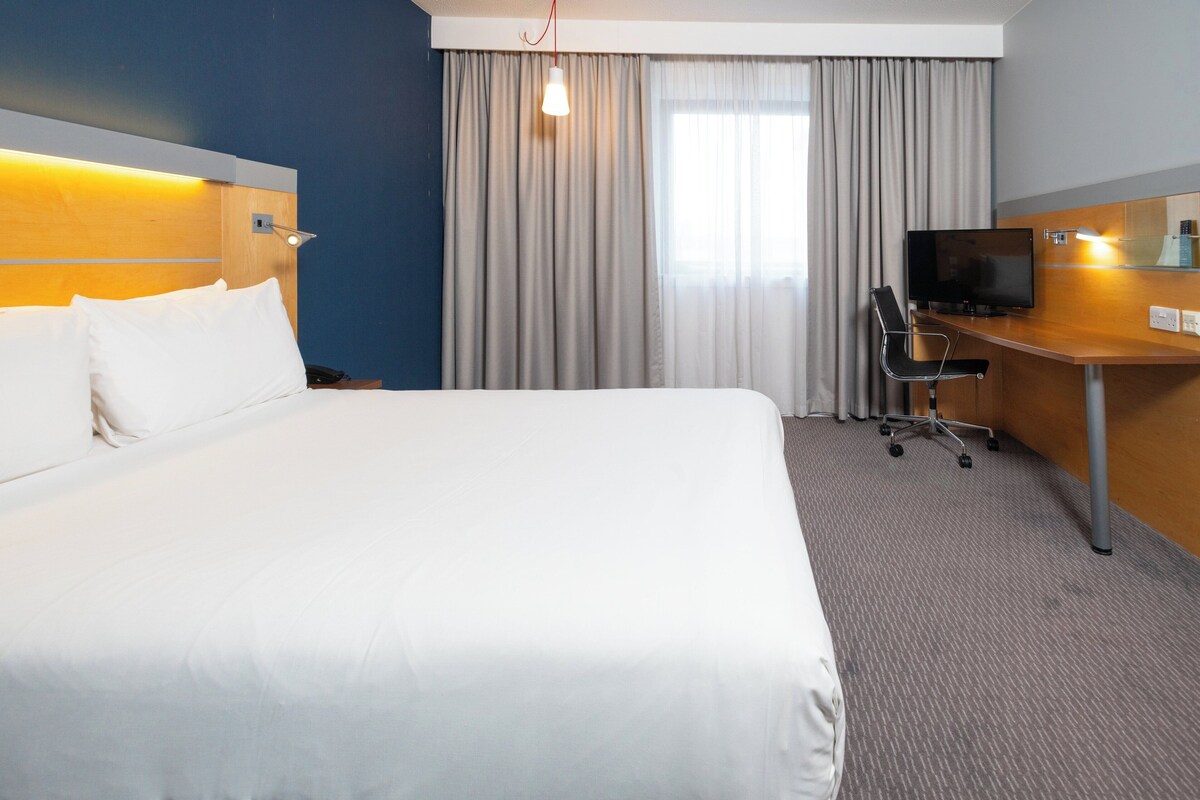 A hotel room with a large bed, a desk with a chair, a television, and a window with curtains.