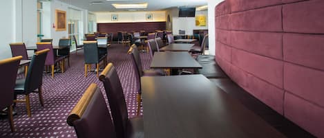 A restaurant with multiple tables and chairs, a patterned carpet, and a red cushioned wall.