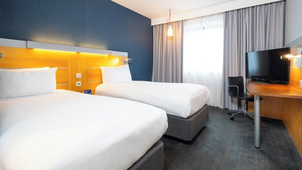 Et hotelværelse med to senge, et skrivebord med tv og et stort vindue med gardiner.